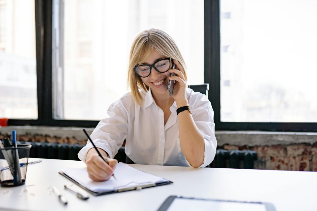 pexels-photo-7793187-7793187 Smiling woman talking on the phone while writing notes at a desk in a modern office.